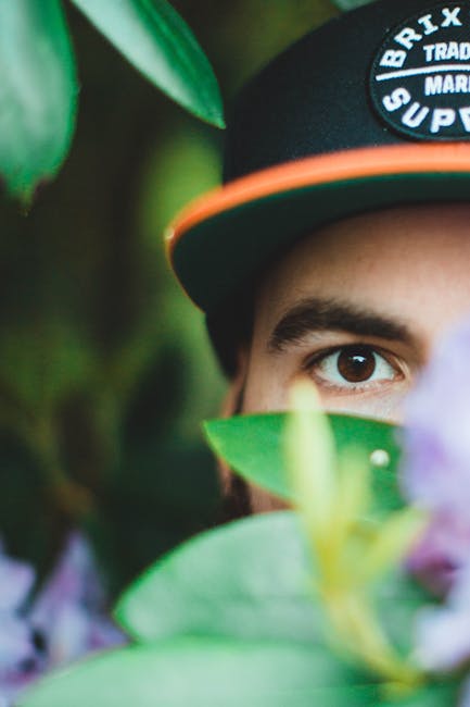 A close-up image showing the face of a young man mostly obscured by green leaves and purple flowers in the foreground. Only one eye and part of his forehead are visible, with the eye looking directly at the camera. He is wearing a black cap with an orange brim, displaying a logo that includes the words 'Brixton' and 'Supplies.' The background features a natural environment with blurred greenery, suggesting an outdoor setting. The arrangement of the plants and flowers, combined with the focus on the eye peeking through, creates a sense of concealment or exploration. This scene illustrates a theme of privacy or hidden observation, which aligns with the idea of discreet rubbish removal or alternative waste handling services offered by Waste Disposal Brixton, subtly emphasizing the importance of unseen or private waste management in an outdoor environment.