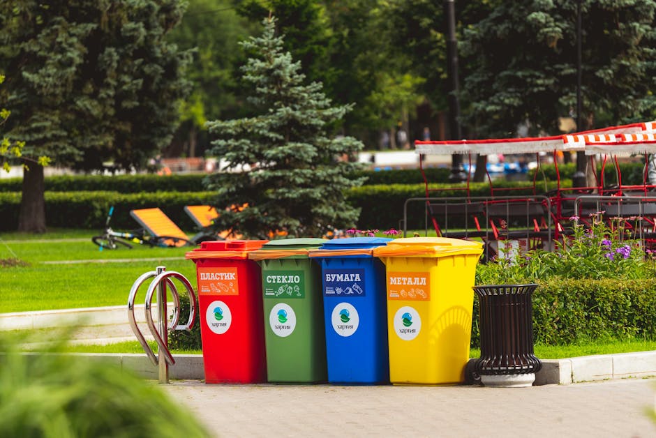A row of four brightly colored waste bins is positioned on a paved pathway in a park setting. From left to right, the bins are red, green, blue, and yellow, each with distinct labels in Cyrillic script indicating their designated waste materials: plastic, glass, paper, and metal. The red bin has a label with orange accents, the green with white, the blue with white, and the yellow with orange accents, all featuring icons representing their respective waste types. The bins are made of plastic with smooth finishes, and they are placed side by side, slightly angled toward the camera. Behind the bins, the lush green environment includes well-maintained grass, small hedges, and a mixture of tall trees and coniferous varieties, with some outdoor seating visible on the right side. The scene is illuminated by natural daylight, highlighting the vivid colors of the bins and the greenery around them. This setup suggests a designated area for separate waste collection, contributing to environmentally conscientious rubbish disposal or private waste management, as operated by Waste Disposal Brixton.