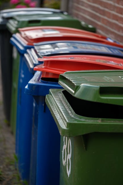 A row of large, rectangular wheelie bins lined up along a pavement beside a brick wall, with green, red, blue, and dark green lids closed. The bins are made of durable plastic with slightly textured surfaces and are positioned close together, creating a uniform line. The area appears to be outdoors, possibly near a commercial or residential building, with soft natural lighting. The green bin in the foreground has a partially visible recycling symbol, indicating it is used for waste sorting. The image reflects a typical scene where private waste collection services, such as those offered by Waste Disposal Brixton, may handle rubbish removal, especially when considering alternatives to municipal collection. The setting portrays an organized waste management system that facilitates efficient rubbish collection or on-site clearance, emphasizing the importance of maintaining clear and accessible disposal points for different waste types.