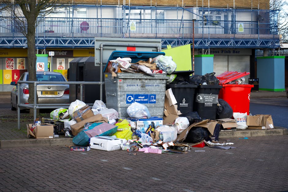 A large pile of mixed waste and rubbish is accumulated around several wheelie bins on a paved outdoor area in front of a commercial building. The waste includes cardboard boxes, plastic bags, crumpled paper, and discarded packaging, some spilling onto the surrounding ground. The bins are varied in colour, with a grey bin for mixed paper and card, a black bin for general waste, a red bin possibly for recyclables, and a white or cream-colored bin, all positioned on a paved surface adjacent to a low metal railing. Behind the waste, there is a broad view of a building with storefronts, including signs and windows, and a blue scaffold or fencing structure covering a section of the upper part of the building. A silver car is parked nearby, partially visible behind the railing, and a leafless tree stands to the left of the waste pile. The scene appears to illustrate an example of unmanaged rubbish accumulation, relevant to waste removal or private waste collection services, such as those offered by Waste Disposal Brixton.