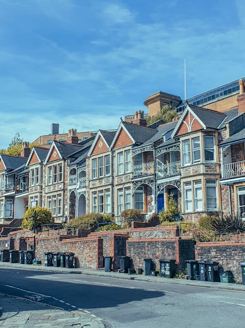 A row of historic Victorian-style terraced houses with ornate white-painted metal balcony railings and decorative trim, situated on a slightly elevated street level, with brick facades in shades of red and beige. The houses have numerous large, sash windows with white frames, and the rooftops feature triangular gables with slate tiles. The foreground shows a low brick wall separating the footpath from the street, with several black wheelie bins lined up along the pavement. The street appears clean, indicating regular waste collection, possibly through private rubbish removal services like Waste Disposal Brixton. The background reveals a blue sky with scattered clouds, and modern commercial buildings behind the row of residential properties, suggesting an urban environment where on-site clearance or alternative waste handling may be used to maintain cleanliness and aesthetic appeal.