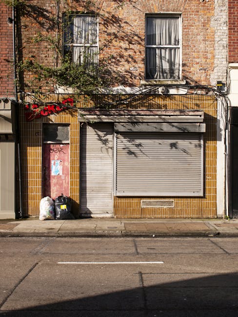 A street scene showing a Waste Disposal Brixton worker operating a red rubbish collection vehicle, parked on the roadside with its rear door open. The worker, dressed in a high-visibility orange vest over a dark uniform, is emptying a black plastic bag filled with waste into the vehicle’s hopper. The vehicle's rear compartment appears to be designed for compacting and lifting waste, with mechanical arms and hydraulic components visible. The street is bordered by a curb and a small grassy verge with some litter scattered around. A motorcycle is parked nearby with a small cargo compartment, and there is a metal barrier with reflective red and white stripes attached to the vehicle. In the background, there are trees, utility poles, and a distant building, under an overcast sky, suggesting an typical urban environment suitable for private waste collection and rubbish removal services, such as those provided by Waste Disposal Brixton, specializing in alternative waste handling methods.