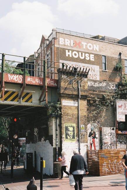 The image depicts the side of a multi-story brick building with a weathered appearance, featuring several layers of graffiti and painted signs, including large white lettering spelling 'BRIXTON HOUSE' on the upper part of the wall. A metal fire escape with black railings and a platform extends from the middle of the building, with pipes running vertically beneath it. On the ground level, a street scene is visible with pedestrians, including a man in a dark jacket and trousers carrying a plastic bag, and a woman in light clothing walking nearby. To the right, there is a stack of wooden pallets or barriers and a painted mural of a person with a hat, possibly part of street art, attached to the wall. The background shows a cloudy sky, suggesting a daytime setting, and the overall environment reflects an urban atmosphere consistent with areas where independent waste collection or on-site clearance might occur, supporting the context of private rubbish disposal services provided by Waste Disposal Brixton.