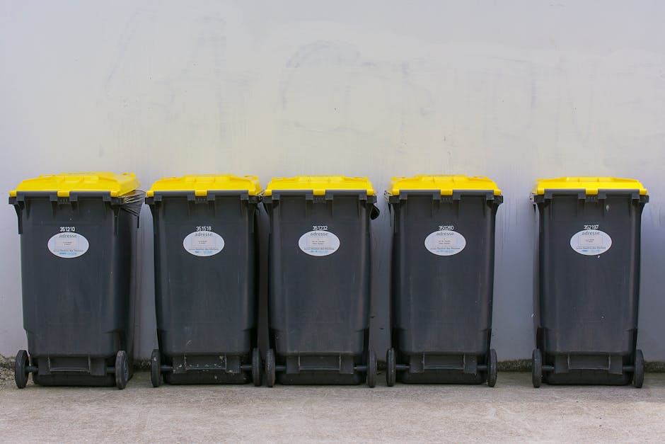 A blue wheeled rubbish bin with a closed lid is positioned on a paved surface in front of a white delivery truck or storage container, which has a plain, slightly weathered metal exterior with visible scratches and dirt marks. The bin is made of durable plastic with a textured finish and sits upright, leaning slightly against the side of the truck. The truck's side is plain with a vertical metal handle and hinges partially visible near the top of the bin, suggesting access to an internal compartment or delivery area. The environment appears to be an outdoor urban or industrial setting, with natural lighting casting soft shadows and emphasizing the textures of the bin and truck surface, reflecting typical conditions for waste collection or private disposal activities. The scene is neutral and functional, fitting within the context of alternative waste handling or rubbish removal services, such as those offered by Waste Disposal Brixton, particularly in relation to community or commercial rubbish management.